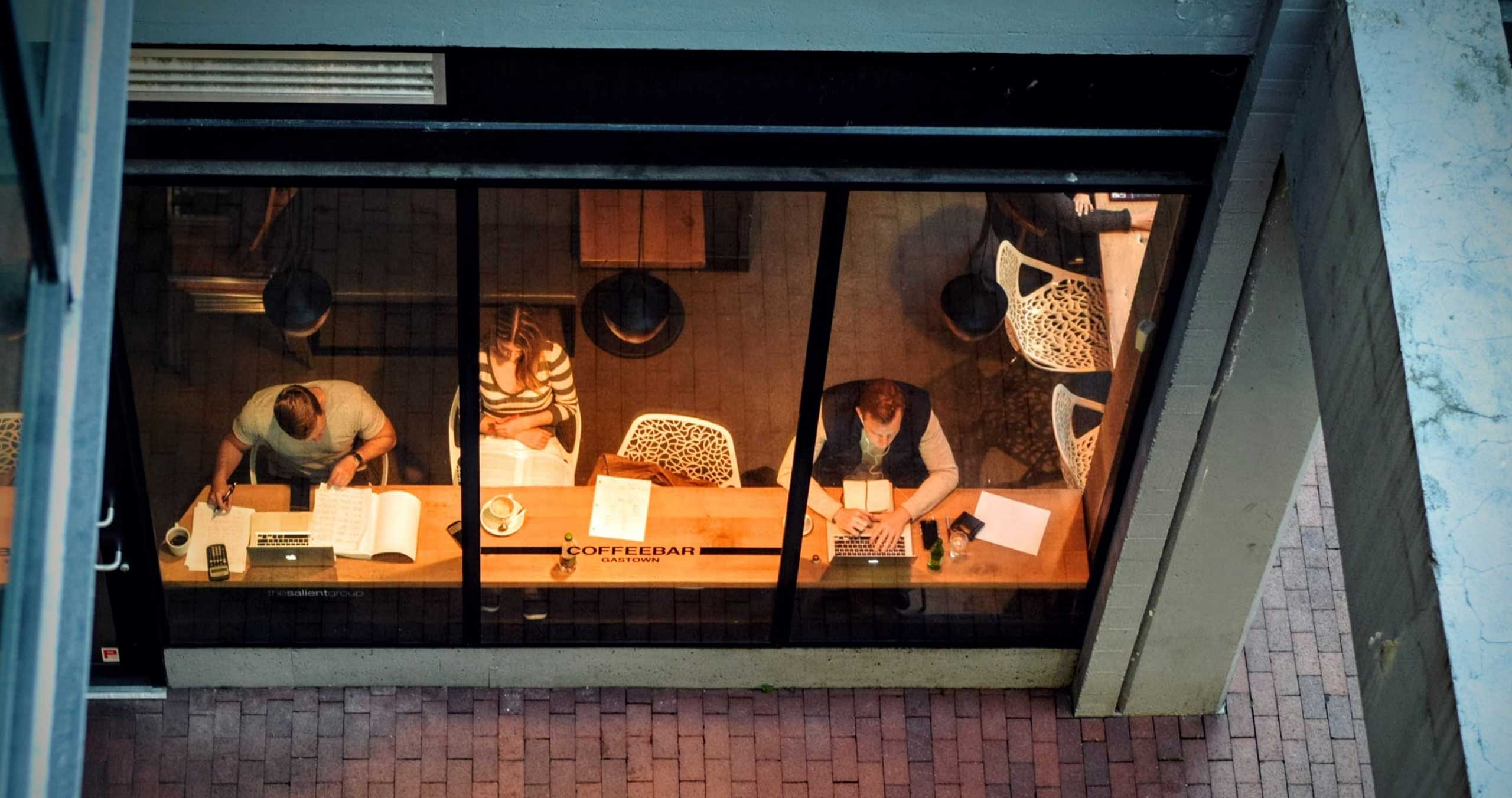 Three people sitting in a cafe working on laptops. The image is taken from above through the window.